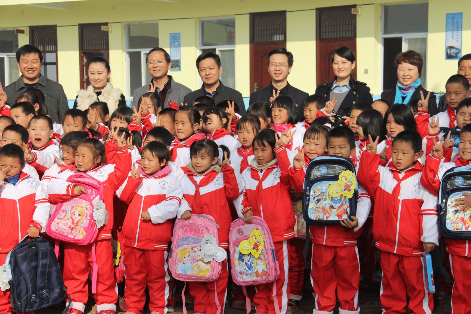 Students in red-and-white uniforms holding new backpacks, gathered with visitors outside a Kangli Hope Primary School