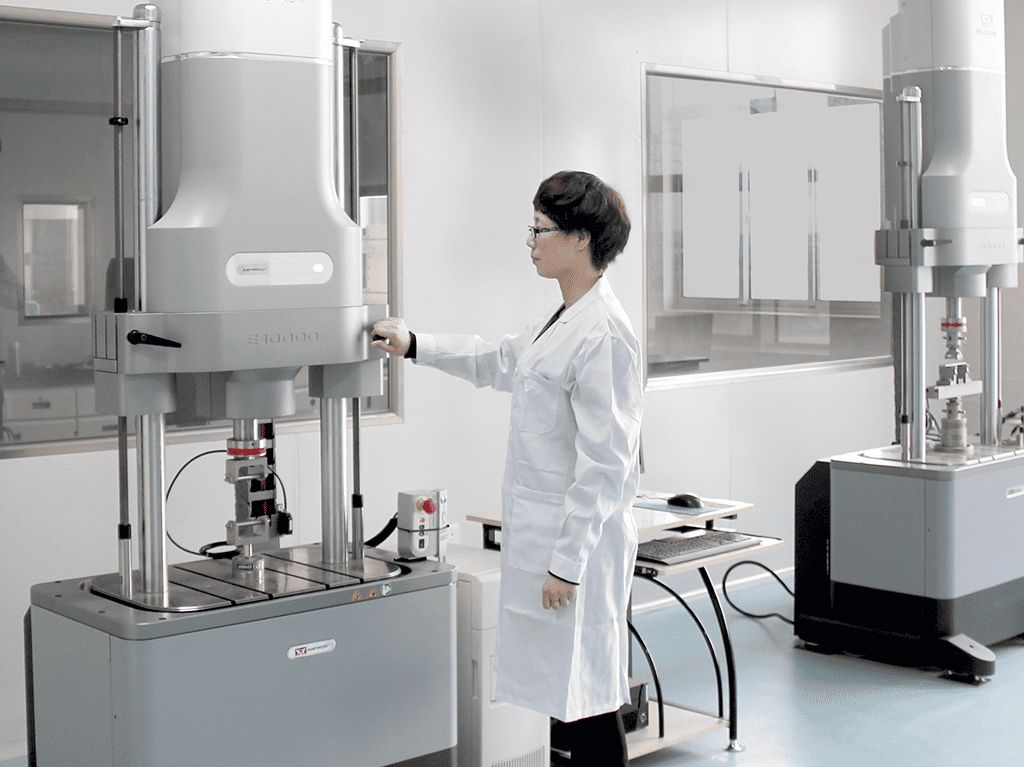 A Kangli technician in a white lab coat operating a servo-hydraulic metal fatigue testing machine in the materials testing laboratory