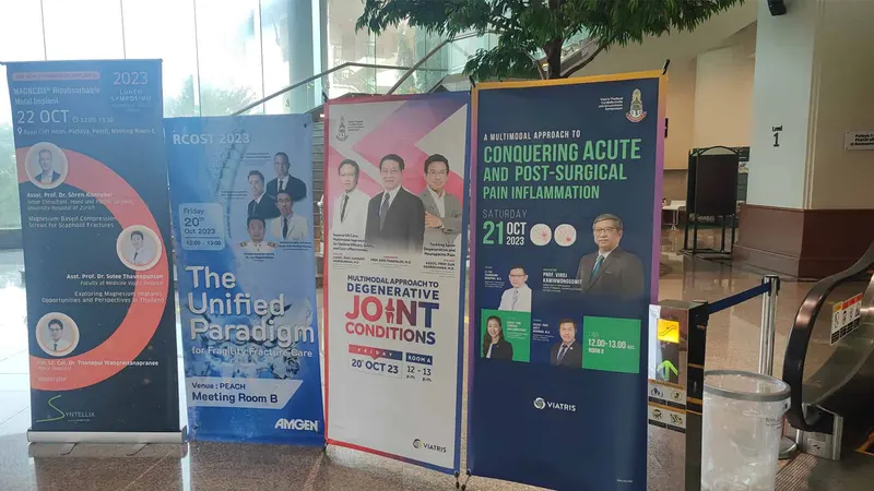 Four freestanding conference banners with speaker portraits beside an escalator in a glass-walled lobby.