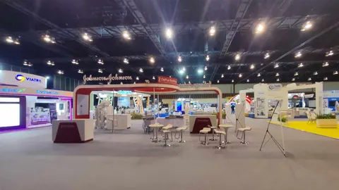 Wide exhibition hall with trade-show booths, illuminated fascia signs, display counters, presentation screens, visitor seating and about six people in the background.