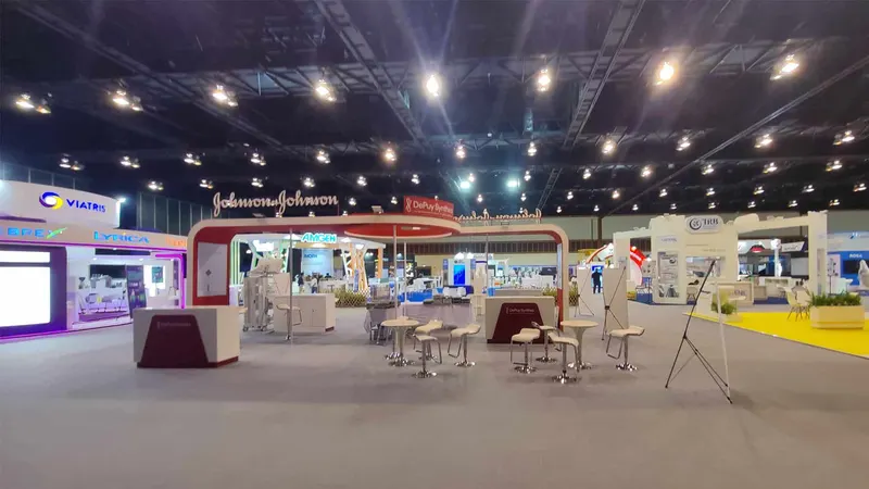 Wide exhibition hall with trade-show booths, illuminated fascia signs, display counters, presentation screens, visitor seating and about six people in the background.