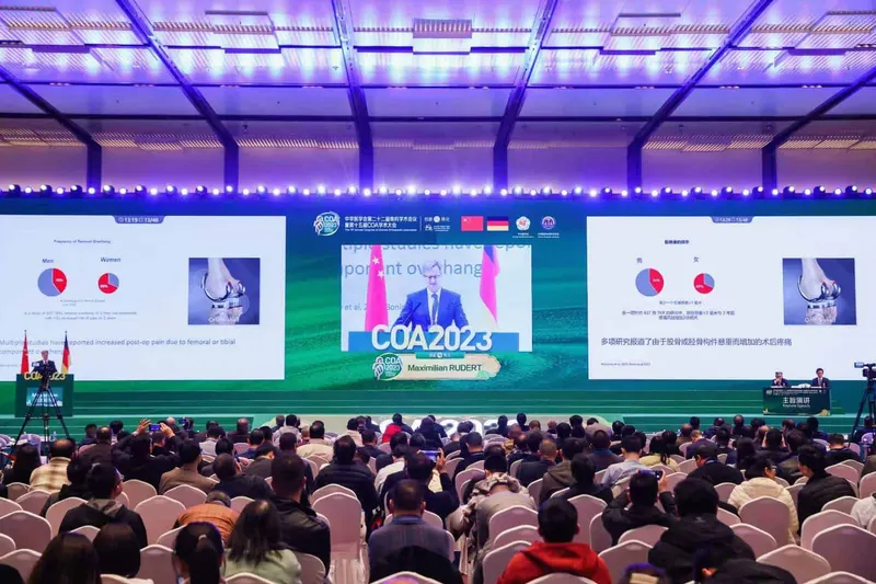 Dozens of attendees sit in rows facing an illuminated conference stage with presentation screens, podiums and a speaker on the central screen.