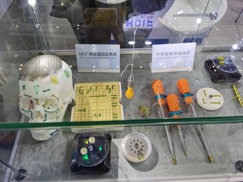 A white skull model with a silver woven mesh and small coloured components sits beside orthopaedic parts in a glass case.