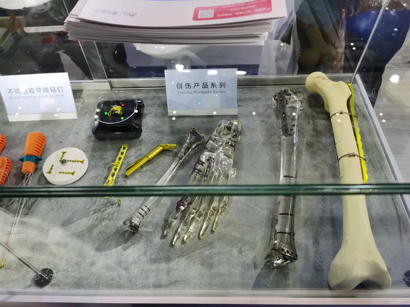 A glass display case on grey fabric holds orange-and-blue instruments, a black component case, yellow parts and gold-toned trauma instruments.