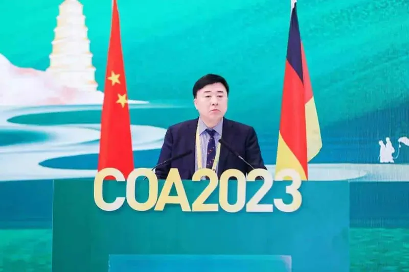 One speaker stands at a turquoise lectern between two flags, with COA2023 lettering across the front.