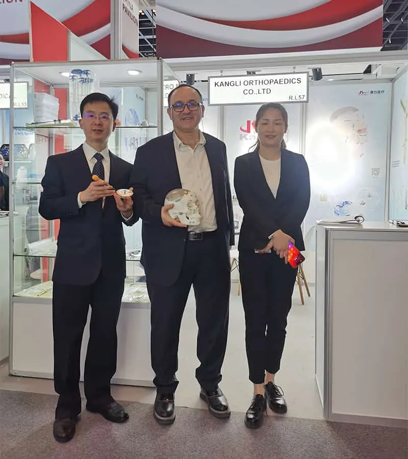 Three participants pose at an exhibition stand holding an orange-handled item, a white anatomical model and a small red-and-blue component.