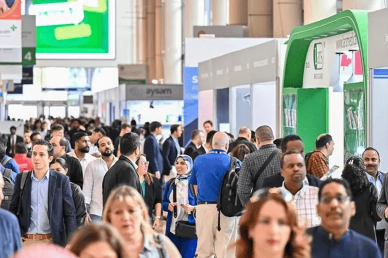 Crowded exhibition aisle with temporary stands, suspended screens and visitors walking or talking beside a large green-and-white booth.