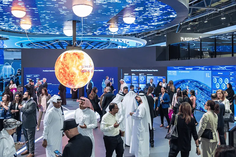 Crowded technology exhibition hall with illuminated display structures, presentation screens and visitors moving between stands.