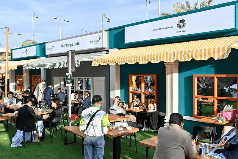 Outdoor exhibition hospitality area with branded façades, café tables, food and groups of visitors dining and talking beneath awnings.