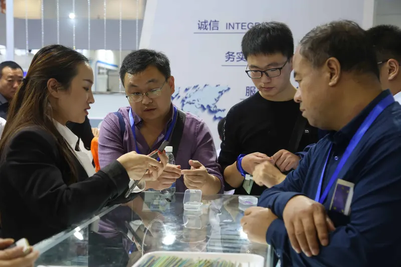 Indoor exhibition stand with six participants gathered around a glass display counter, including people examining a small item and discussing it.