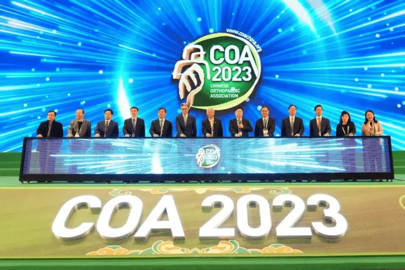 Thirteen formally dressed participants sit behind a long stage desk beneath a blue LED backdrop marked “COA 2023”; several wear lanyards and badges.