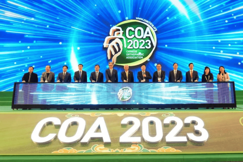 Thirteen formally dressed participants sit behind a long stage desk beneath a blue LED backdrop marked “COA 2023”; several wear lanyards and badges.