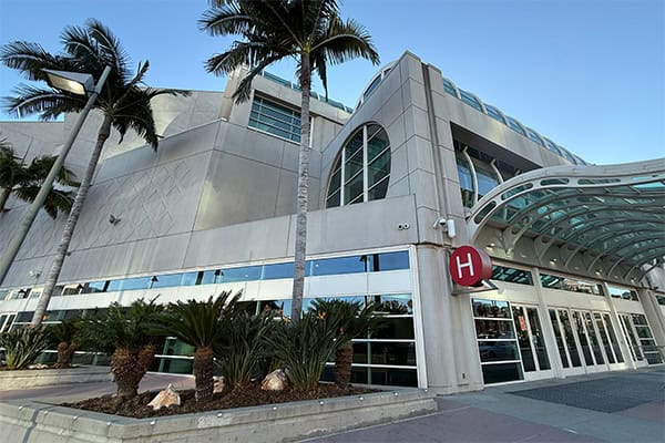 A modern pale-gray and blue-green building exterior with palm trees, reflective windows, and a curved entrance canopy.