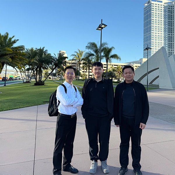 Three people pose on an outdoor plaza beside a large modern building with palm trees.