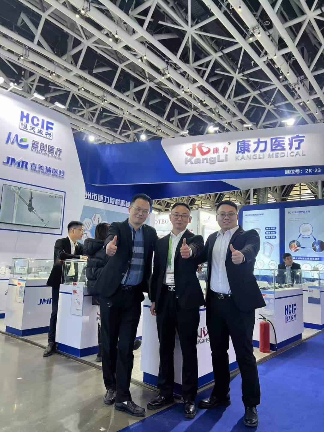 Six participants stand at a trade-exhibition stand, with three in front on blue carpet giving thumbs-up gestures and three partly visible behind the displays.