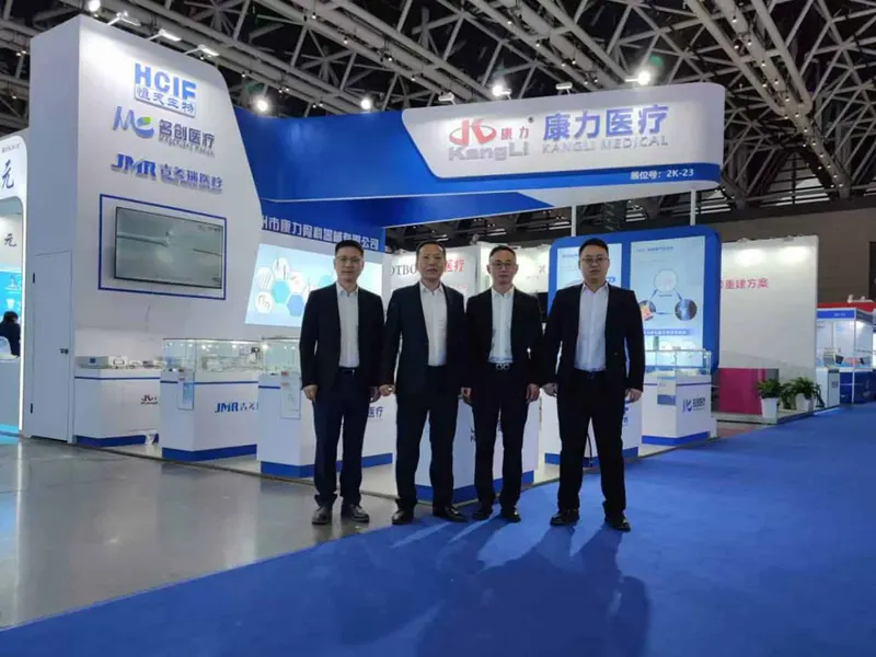 Four people in dark business suits stand in a row before a blue-and-white exhibition stand with glass counters and illuminated screens.