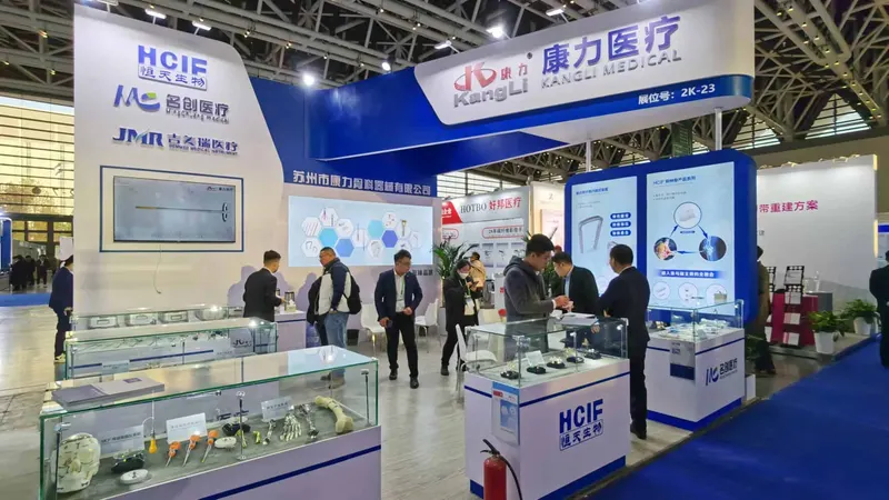 About ten attendees gather around a blue-and-white exhibition stand with glass display cases, information screens and overhead signs reading KANGLI MEDICAL.
