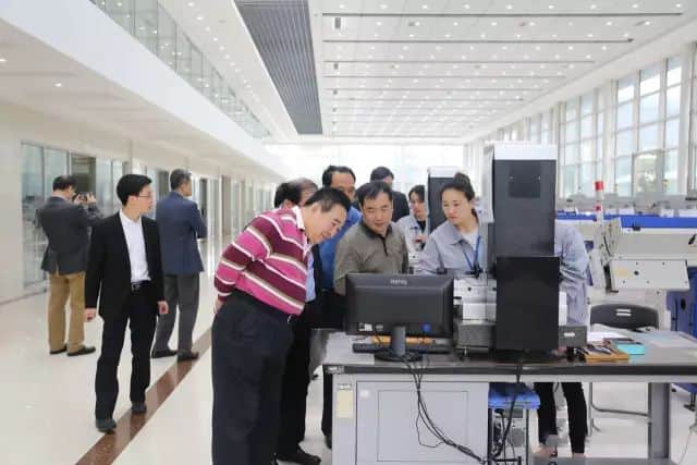About twelve people gather around a computer workstation and industrial equipment in a bright production or laboratory hall.