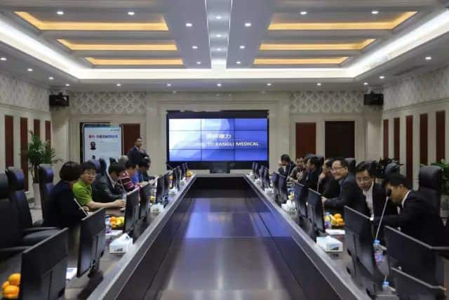 Approximately twenty people sit at conference tables with individual monitors facing a large presentation screen.