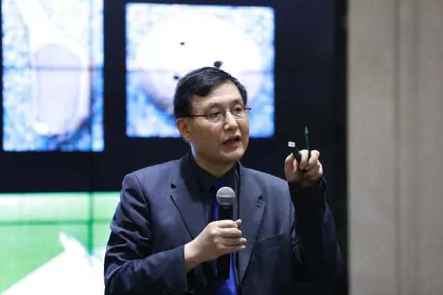 One speaker in a navy jacket and blue tie holds a microphone and presentation pointer before a multi-panel display.