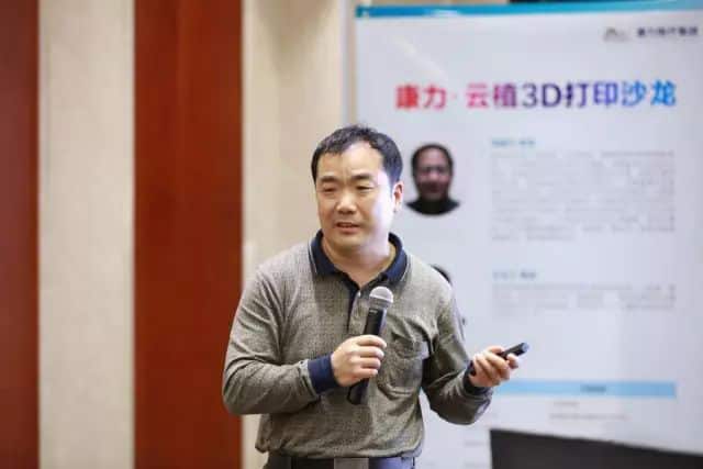 One speaker in a grey sweater holds a microphone and remote before a white-and-blue presentation backdrop.