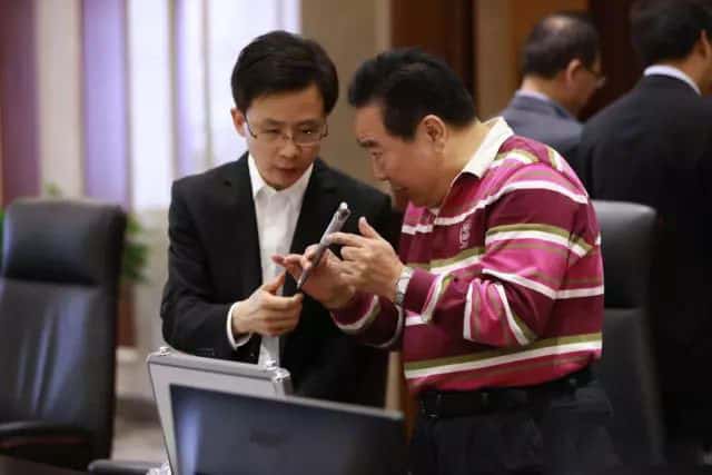 Two people lean over a table and examine a small handheld object, with two partly visible participants in the background.