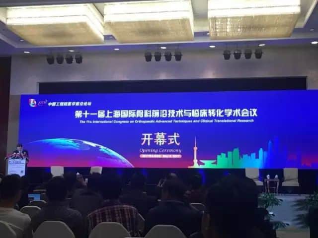 Dozens of seated conference attendees face a stage with a podium and illuminated blue and violet event backdrop.