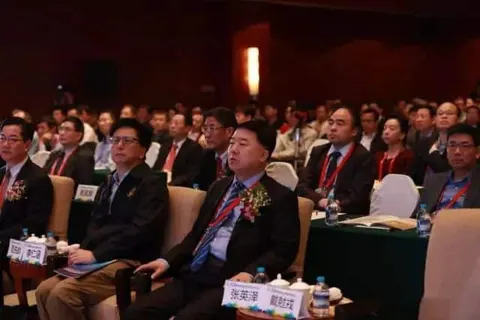 Rows of roughly 50 seated conference attendees face an unseen presentation area at tables with name cards, papers and lanyards.