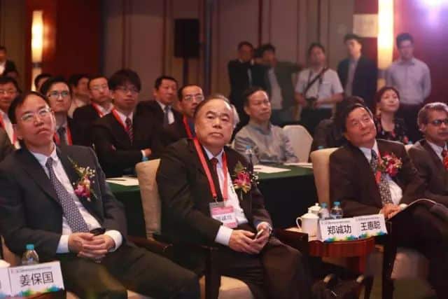 Approximately 20 conference attendees sit in rows wearing red lanyards and name cards, with several standing along the back wall.
