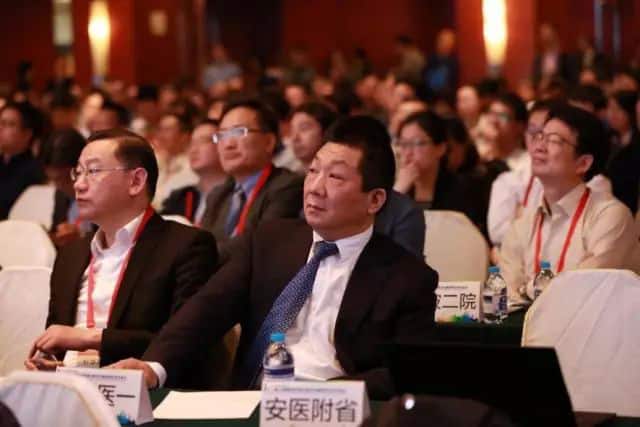 Around 30 seated conference participants in dark jackets and event lanyards face an unseen presentation area.