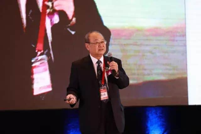 One suited speaker stands on a stage holding a microphone and presentation remote in front of a projected screen.