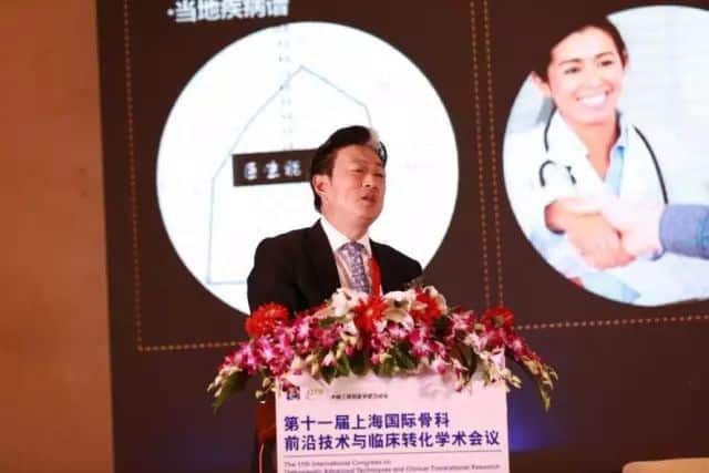 One suited speaker stands behind a flower-decorated podium while projected graphics show a person in a white coat shaking a hand.