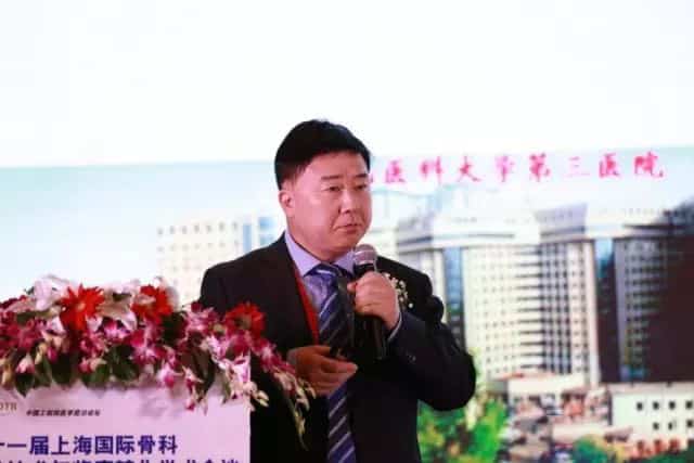 One participant in a dark suit holds a handheld microphone in front of a printed event backdrop and floral arrangement.