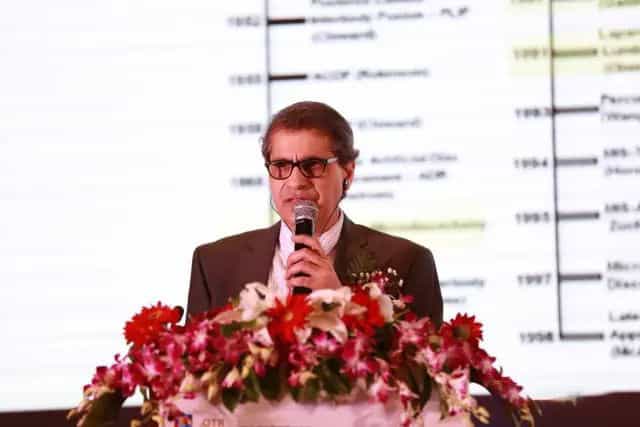 One participant speaks into a handheld microphone behind a flower-decorated lectern with an information slide projected behind.