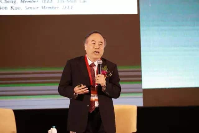 One suited participant speaks into a handheld microphone before projected screens and horizontal illuminated stage bands.