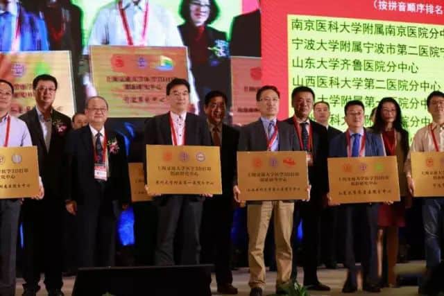 About 14 participants stand on a lit stage holding matching rectangular presentation boards, with a red and black screen behind them.