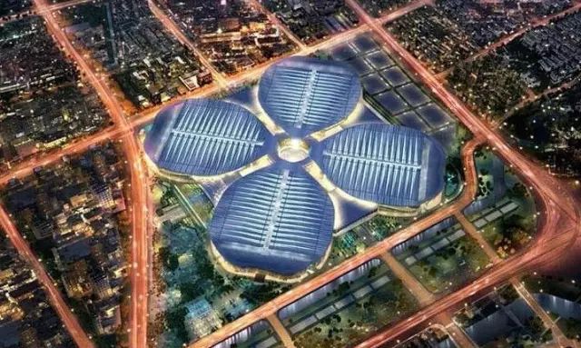 Four blue-gray, ribbed exhibition halls surround a circular central structure in an aerial night view, with illuminated roads and walkways around the complex.