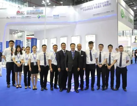 Fourteen participants stand in one row at an indoor medical-equipment exhibition, with a branded booth, display panels, blue carpet and exhibition lighting behind them.