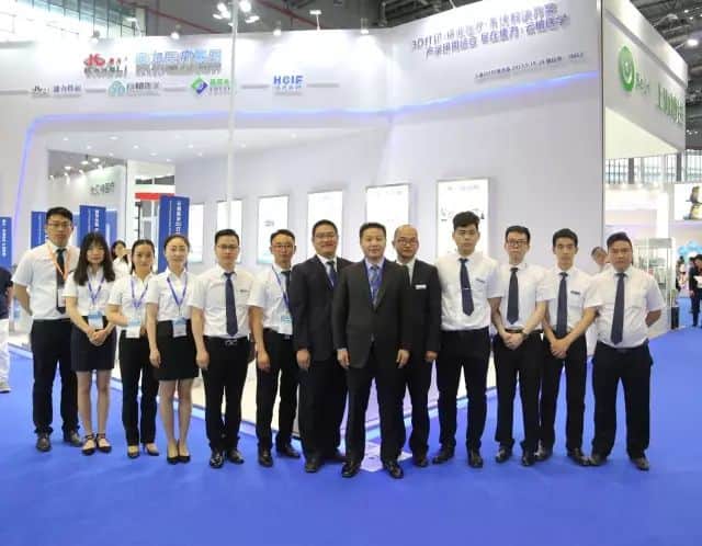 Fourteen participants stand in one row at an indoor medical-equipment exhibition, with a branded booth, display panels, blue carpet and exhibition lighting behind them.