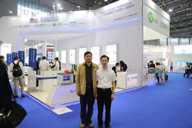 Two participants stand in front of a large illuminated medical-equipment exhibition stand while other visitors move through the carpeted aisle.