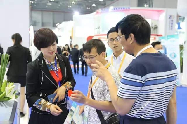 Four participants gather beside display panels in a crowded medical-equipment exhibition hall, with blue-carpeted aisles and other attendees behind them.