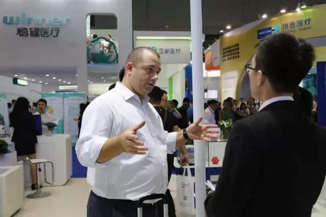 Two participants stand in the foreground of a busy medical-equipment exhibition hall, one gesturing beside a suitcase and the other facing the booth.
