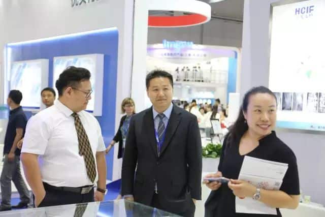 Three participants stand near illuminated display panels at an indoor medical-equipment exhibition stand, with additional visitors behind them.