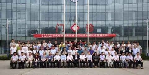Approximately sixty adults stand in rows outdoors in front of a building, with seated participants in the front row, identification lanyards and a red event banner.