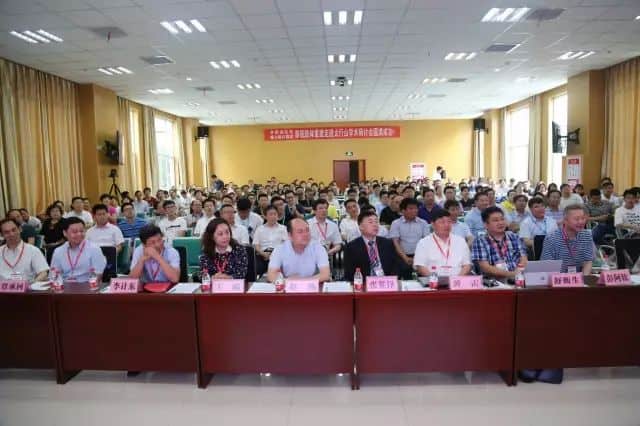 Approximately one hundred adults sit at conference tables in an indoor hall with nameplates, water bottles, papers and a banner across the rear wall.