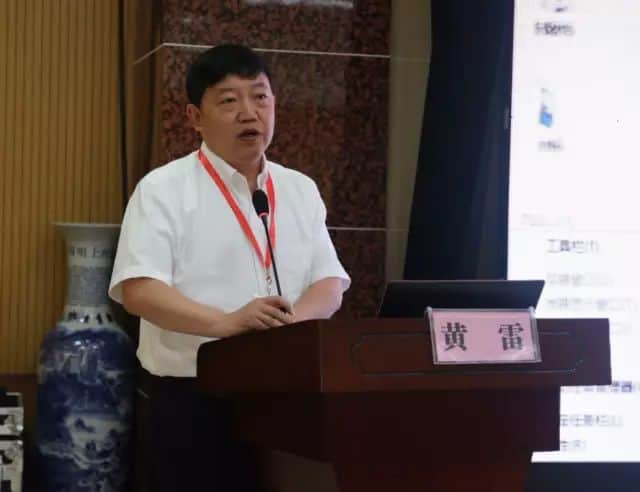 One participant in a white collared shirt stands at a dark wooden lectern with a microphone, while a projected slide appears at the right.