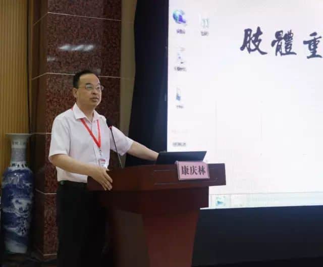 One participant in a white collared shirt stands at a lectern and points toward a large presentation screen beside a laptop.
