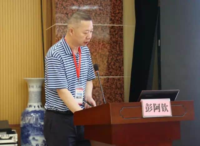 One participant in a striped polo shirt stands at a lectern beside a microphone, laptop and large projection screen.