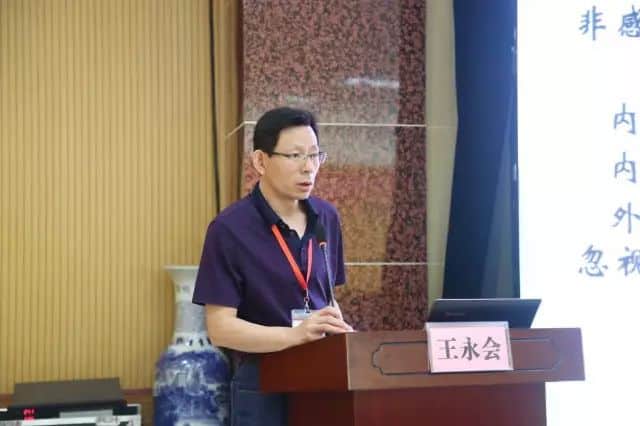 One participant in a dark purple collared shirt and glasses stands at a lectern beside a projected presentation screen and open laptop.