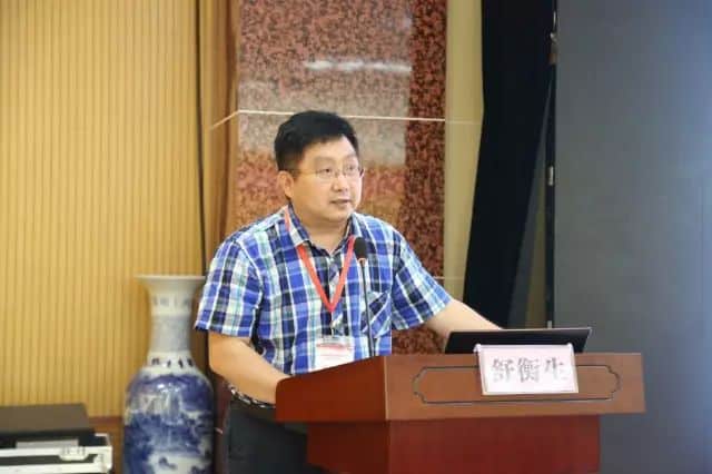 One participant wearing glasses and a plaid shirt stands at a lectern with a microphone and laptop during an indoor presentation.